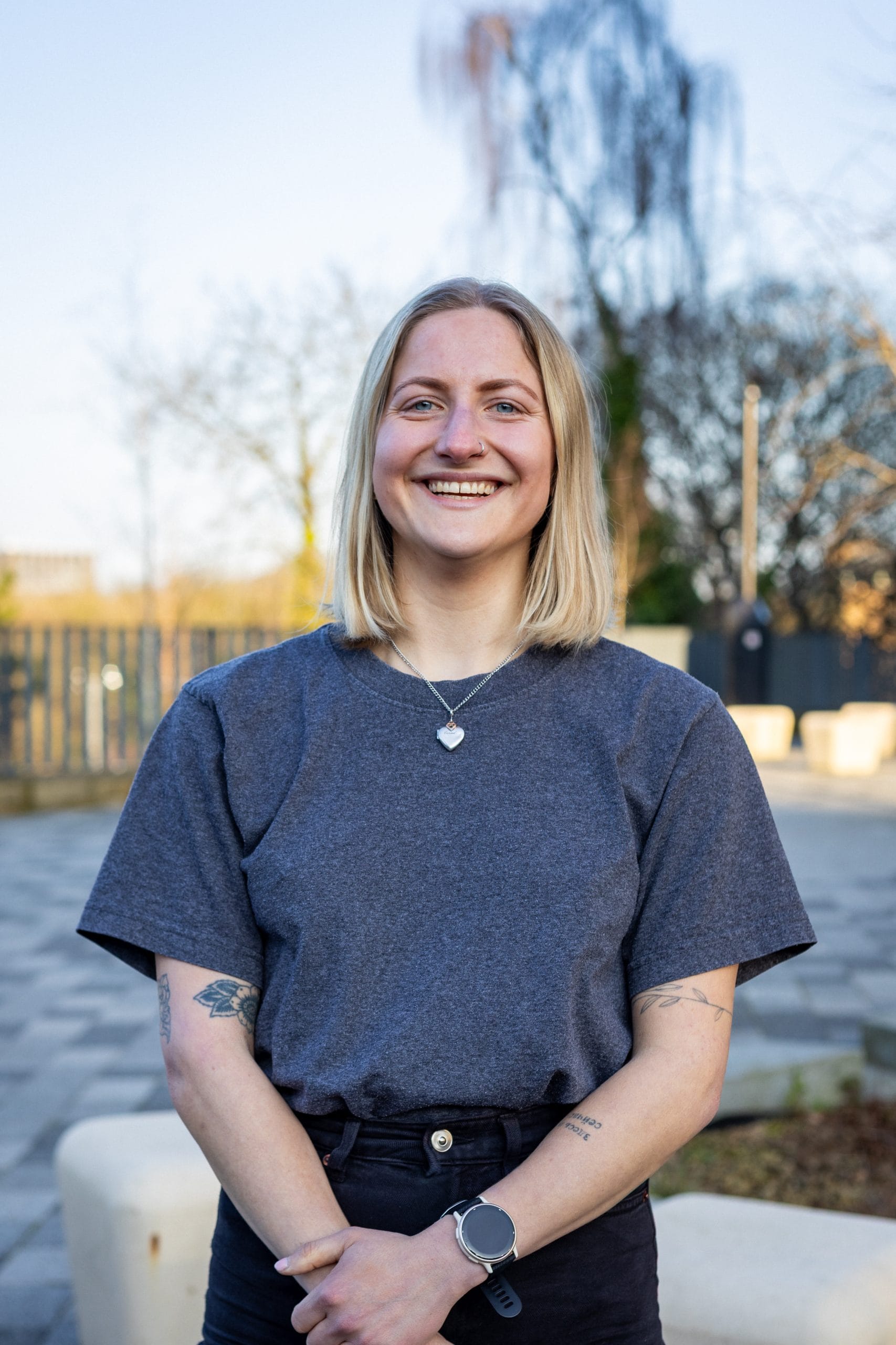 Blonde woman standing outside, smiling at the camera and wearing a grey tshirt