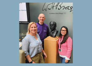 4 people smile in front of a bust of Sir William Henry Bragg