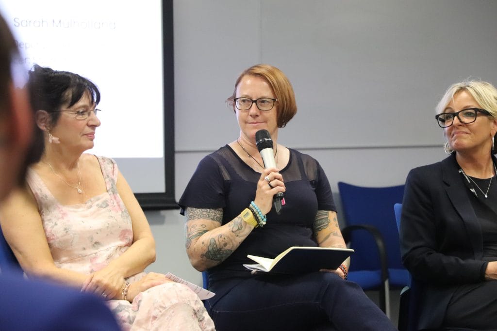 A panel of three women. The women in the centre has a microphone. 