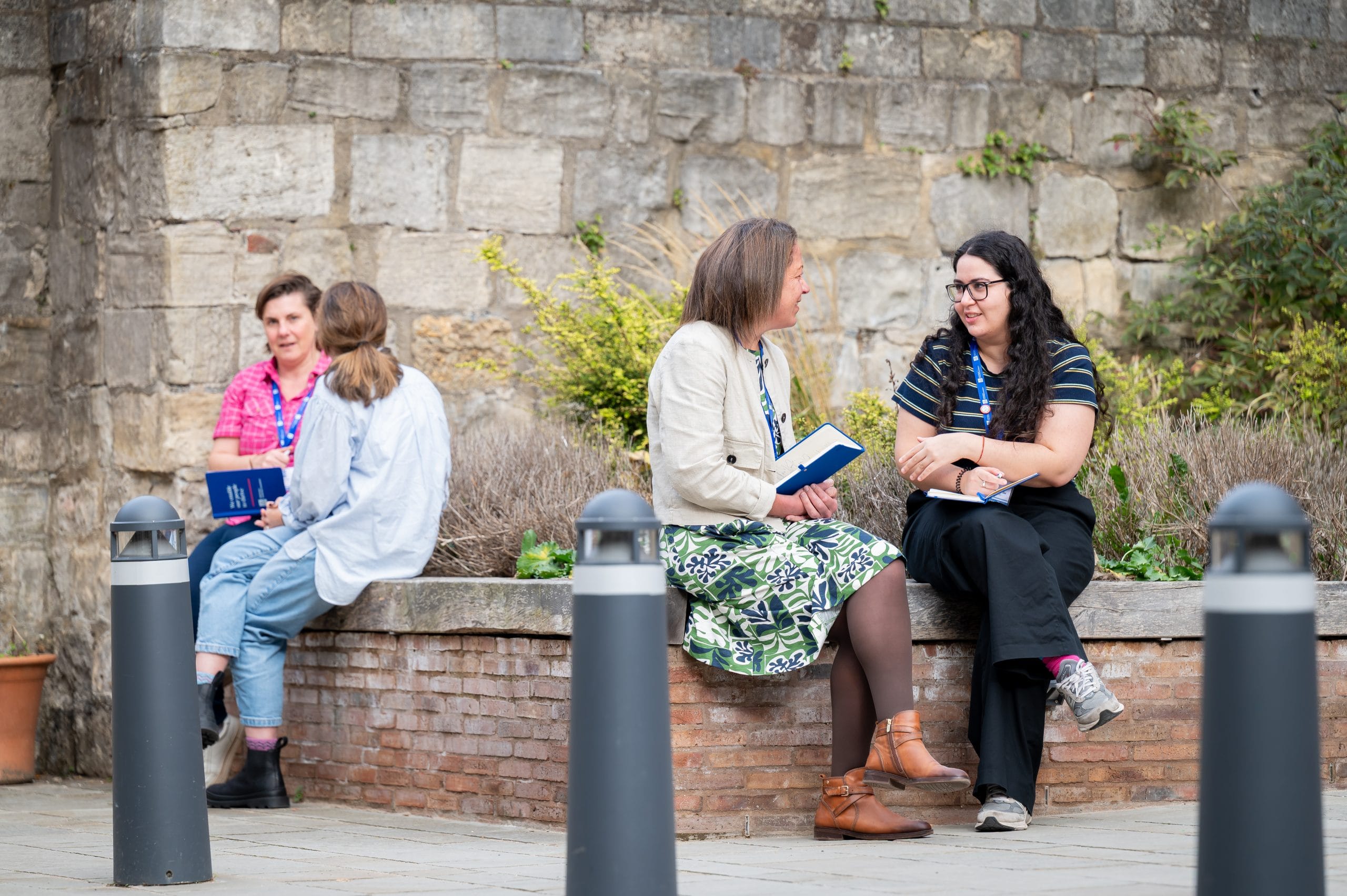 Two women talking and sitting on wall with notebooks.