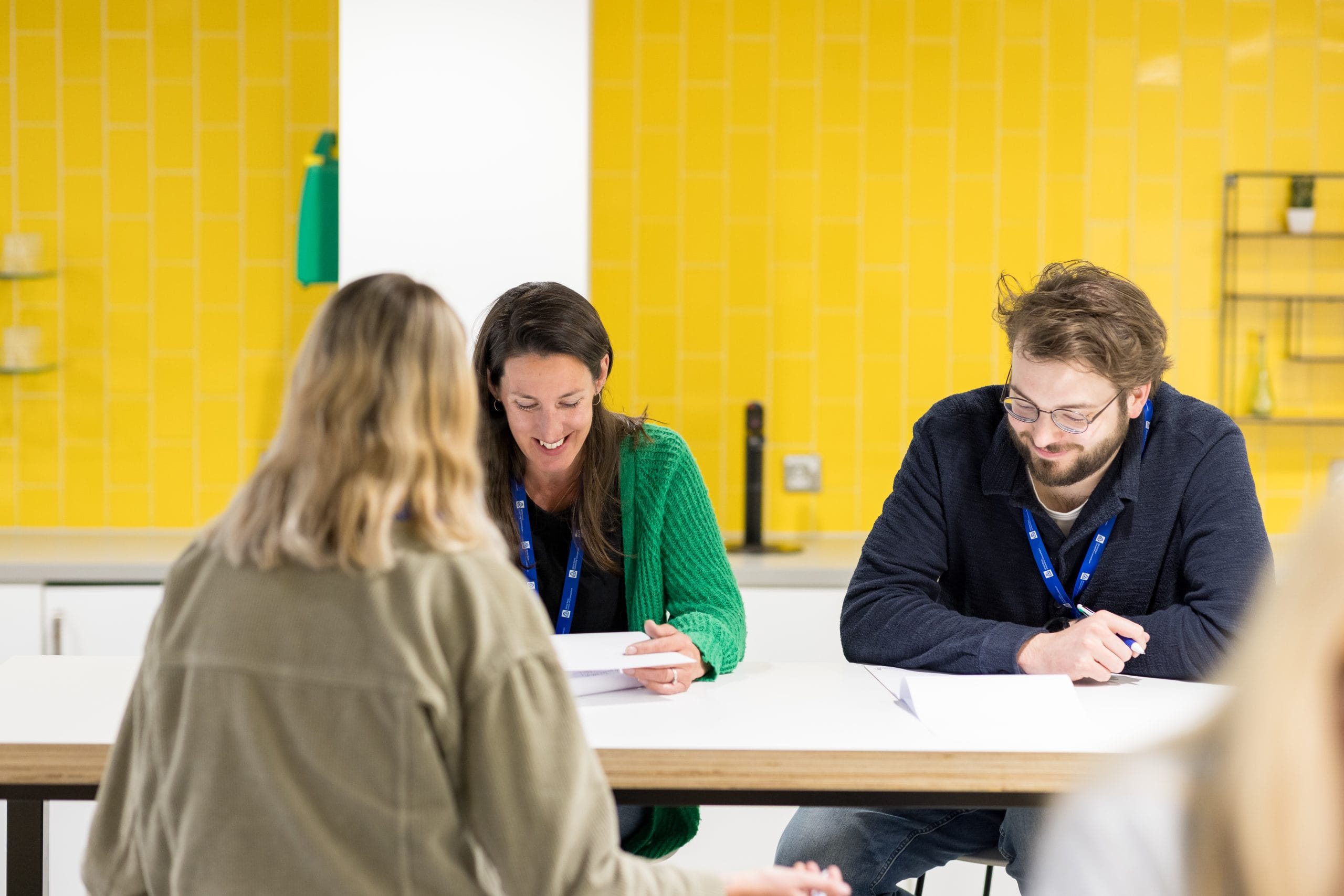 Three people sat at desk with white rose lanyards talking and smiling