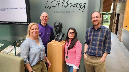 4 people smile in front of a bust of Sir William Henry Bragg